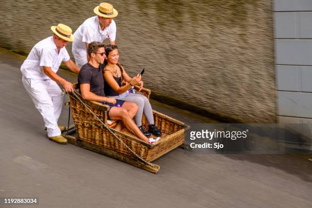 tourists taking a ride in a tradional wicker toboggan down the hill from monte in funchal on madeira island in portugal - madeira material stock pictures, royalty-free photos & images