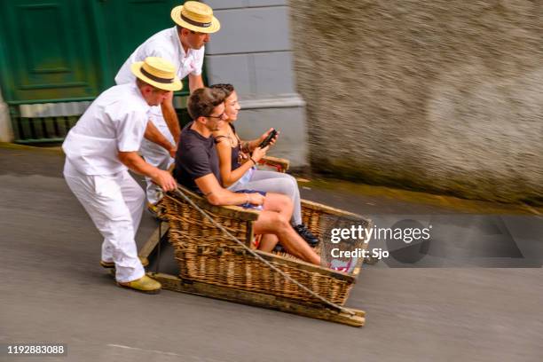 tourists taking a ride in a tradional wicker toboggan down the hill from monte in funchal on madeira island in portugal - monte stock pictures, royalty-free photos & images