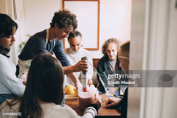 friends looking at teenage boy preparing smoothie at dining table in room - blender stock-fotos und bilder