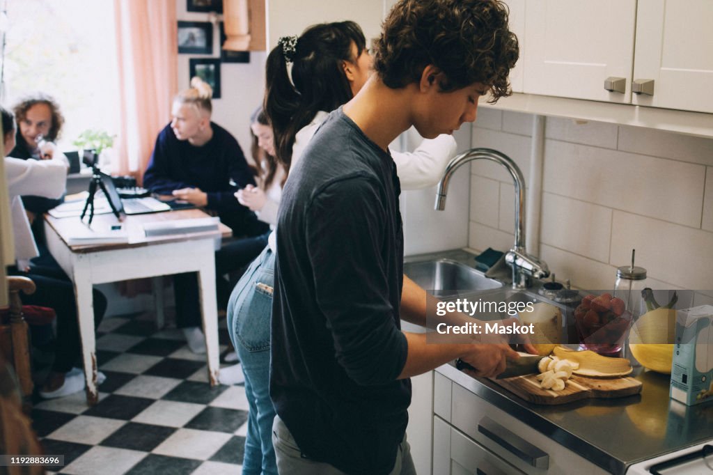 Teenage boy with female cutting fruits at kitchen counter while friends sitting in background