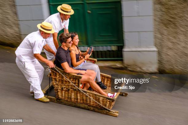 tourists taking a ride in a tradional wicker toboggan down the hill from monte in funchal on madeira island in portugal - monte stock pictures, royalty-free photos & images