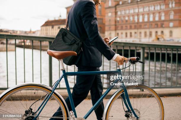 midsection of businessman using smart phone while walking with bicycle on bridge in city - skandinavien stock-fotos und bilder