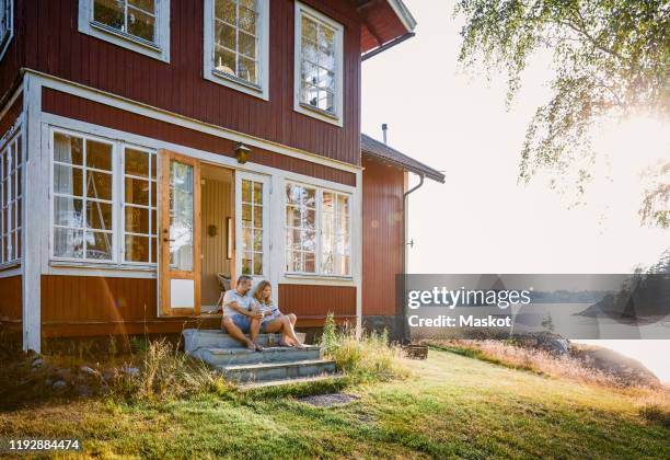 full length of couple sitting at log cabin entrance by lake during summer - cabin scandinavia stock pictures, royalty-free photos & images