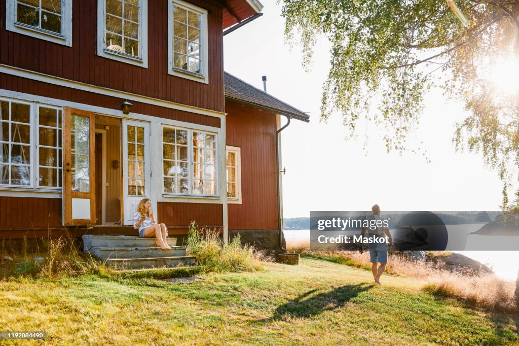 Woman sitting on steps at entrance of log cabin while man walking towards lake on sunny day