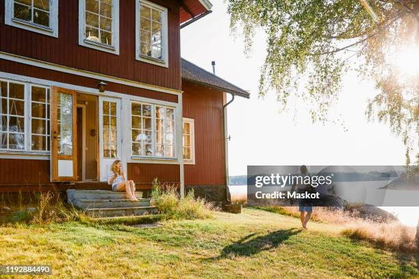 woman sitting on steps at entrance of log cabin while man walking towards lake on sunny day - 40s couple sunny stockfoto's en -beelden