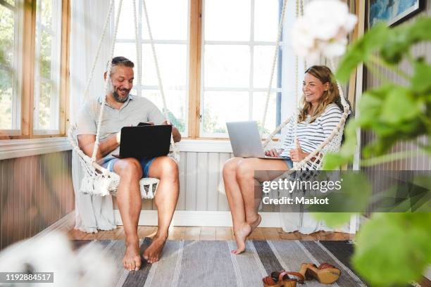 Smiling couple using laptops while sitting on rope swing in log cabin