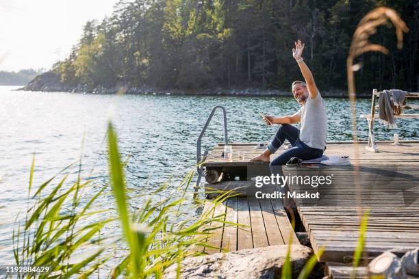 mature man looking away and waving while holding digital tablet on jetty over lake - mann winkt stock-fotos und bilder