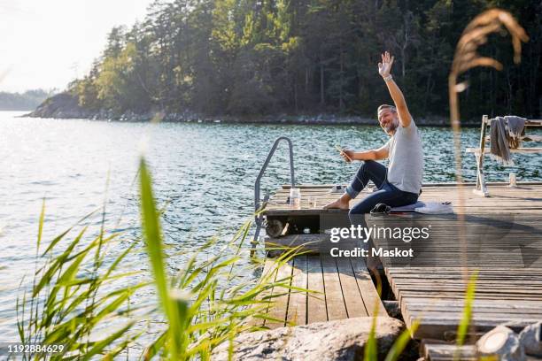 mature man looking away and waving while holding digital tablet on jetty over lake - jetty lake stock pictures, royalty-free photos & images