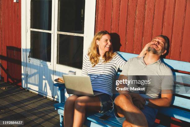 happy woman using laptop while playing with man on bench at porch during summer - cabin stockfoto's en -beelden