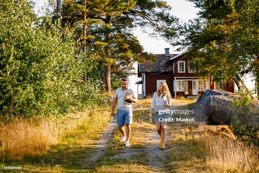 Full length of couple holding laptops while walking on footpath during sunny day