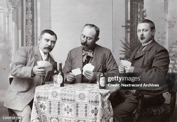 Three men in a card game, gambling, at leisure, while smoking cigars and drinking whiskey, depicted in a vintage cabinet card portrait taken in the...