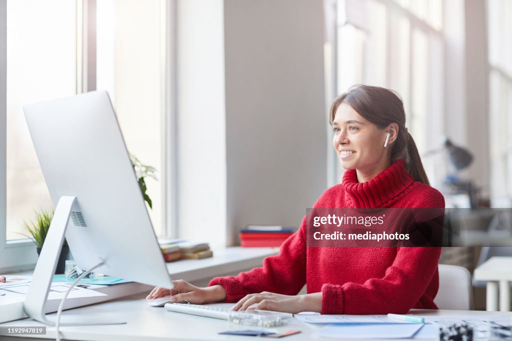Smiling pretty skilled young woman in red knitted sweater and wireless earbuds sitting at table in office and working with modern computer
