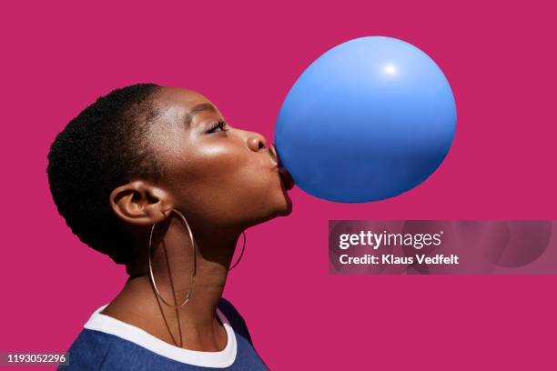 young woman blowing balloon against pink background - fashion photography stock-fotos und bilder