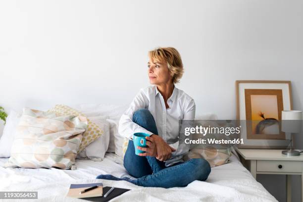 relaxed mature woman sitting on bed at home - white bed cushion stock-fotos und bilder