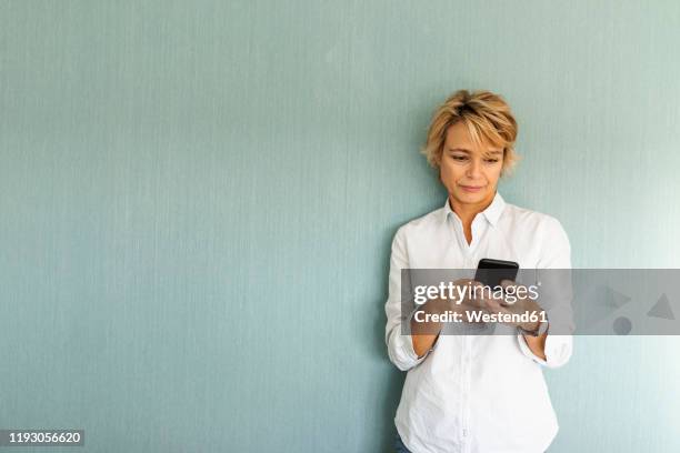 portrait of mature woman standing at a wall using smartphone - woman white shirt stock-fotos und bilder