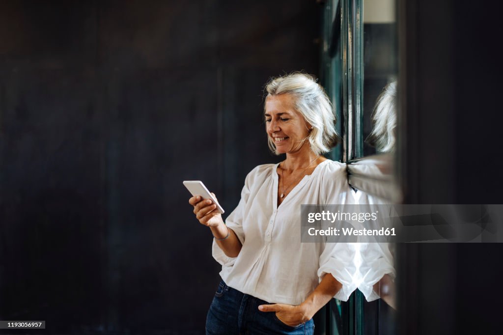 Smiling casual mature businesswoman using smartphone at the window in loft office