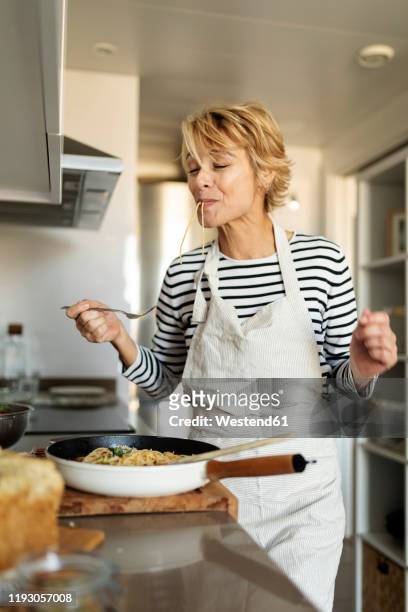 mature woman tasting homemade pasta dish in kitchen at home - kitchen apron stock pictures, royalty-free photos & images