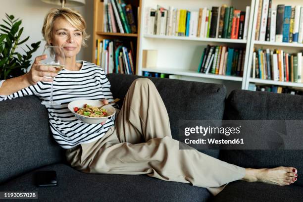 mature woman eating homemade pasta dish and drinking wine on couch at home - mature women eating stock pictures, royalty-free photos & images