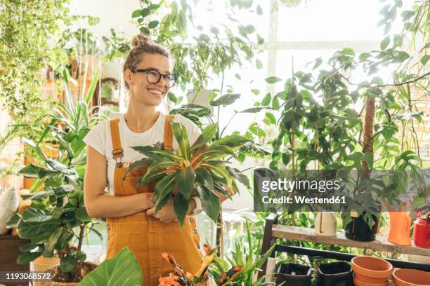 happy young woman holding a plant in a small gardening shop - indoor plant stock pictures, royalty-free photos & images