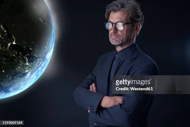 portrait of mature businessman against dark background watching floating planet earth - blauw pak stockfoto's en -beelden