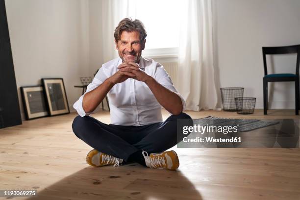 portrait of smiling mature man sitting on the floor at home - gambe incrociate foto e immagini stock