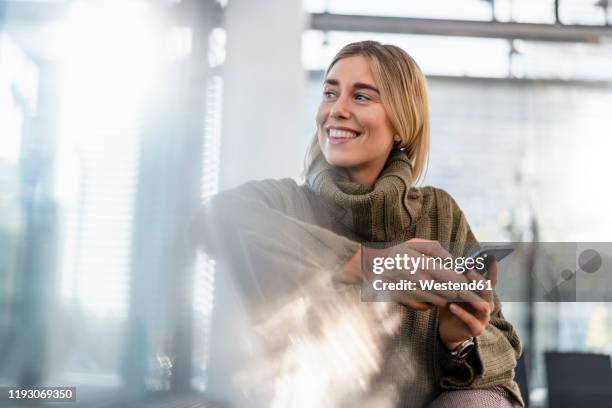 smiling young woman with cell phone sitting in waiting area looking around - on the move fotografías e imágenes de stock