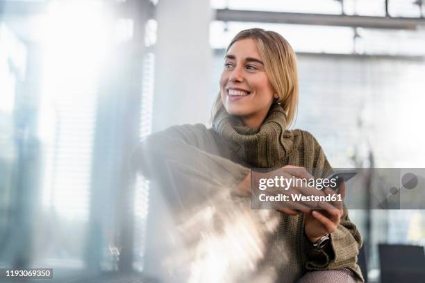smiling young woman with cell phone sitting in waiting area looking around - depth of field imagens e fotografias de stock