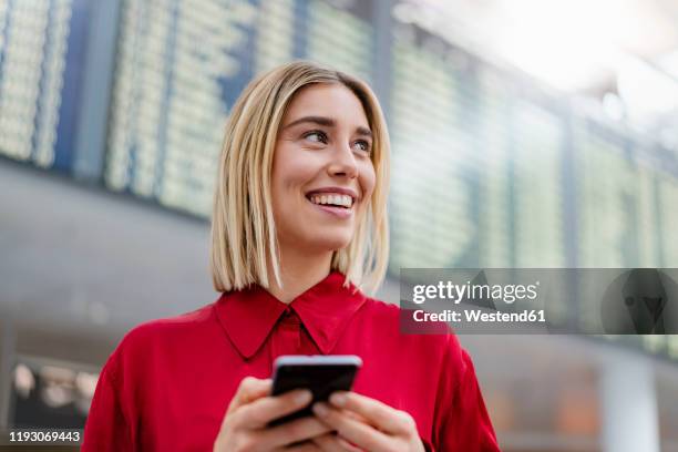 smiling young businesswoman with cell phone at arrival departure board at the airport - women in see through shirts stock pictures, royalty-free photos & images