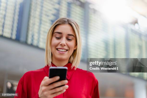 smiling young businesswoman using cell phone at arrival departure board at the airport - departure board front on fotografías e imágenes de stock