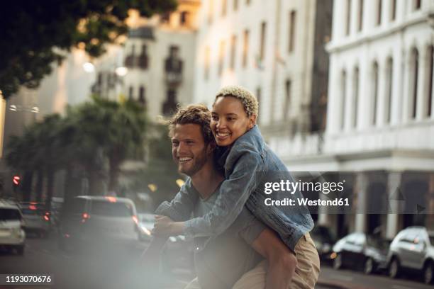 happy young man carrying girlfriend piggyback in the city - piggyback stock pictures, royalty-free photos & images