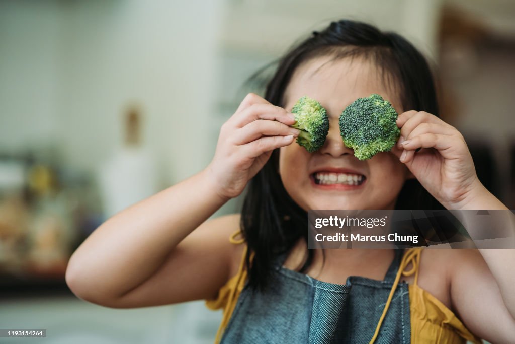 Asiatische chinesische weibliche Kind handeln niedlich mit Hand halten Brokkoli setzen vor ihren Augen mit lächelndem Gesicht in der Küche