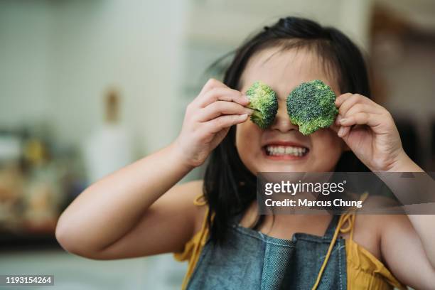 asiatische chinesische weibliche kind handeln niedlich mit hand halten brokkoli setzen vor ihren augen mit lächelndem gesicht in der küche - speisen und getränke stock-fotos und bilder