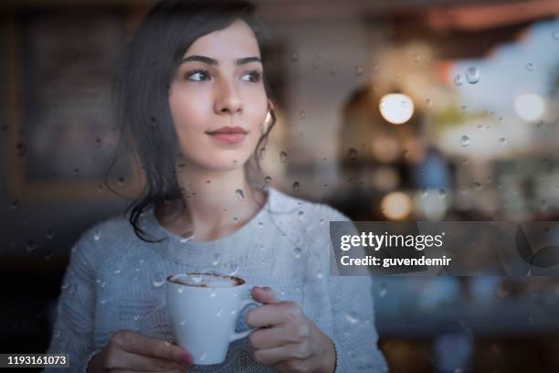 jonge vrouw staande voor raam en genieten van een kopje koffie thuis op een regenachtige dag - at a glance stockfoto's en -beelden