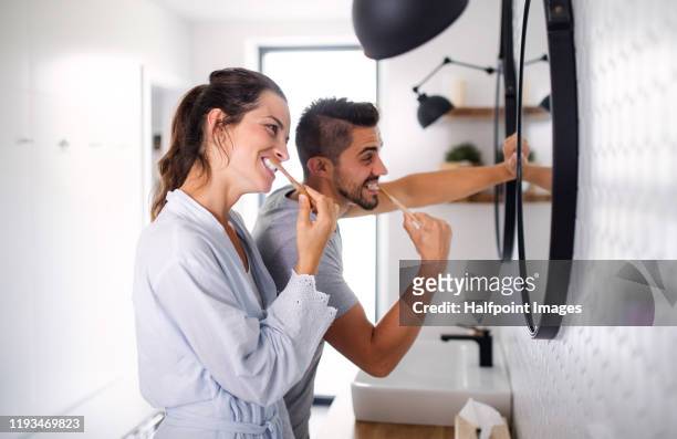 portrait of young couple standing indoors in bathroom at home, brushing teeth. - brush teeth stock pictures, royalty-free photos & images