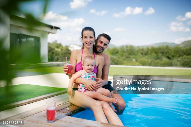 portrait of young couple with toddler girl sitting by swimming pool outdoors at home. - babyschwimmen stock-fotos und bilder