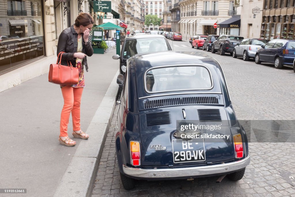 Woman standing beside parked Fiat 500L vintage 1960s car in Parisian street