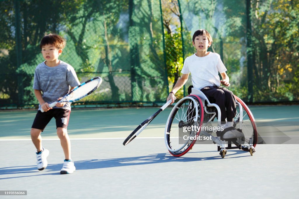 Young boy with his older brother on a wheelchair playing tennis together on a tennis court