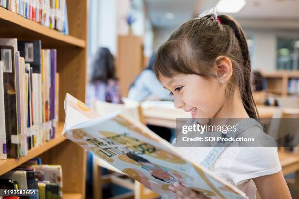 happy little girl reads book in school library - book library stock pictures, royalty-free photos & images