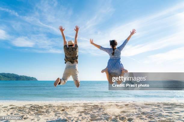 happy asian couple tourists jumping on beach vacations. travel concept of young couple cheering for summer holidays showing success, happiness, and joy on perfect white sand tropical beach under the sun. - phuket beach stock-fotos und bilder