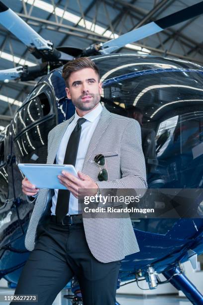 executive man with brown hair and beard dressed in gray suit is leaning on the blue helicopter in which he is going to fly on business theme in his hands he has his tablet people and in his jacket pocket his black glasses - beard pilot stock pictures, royalty-free photos & images