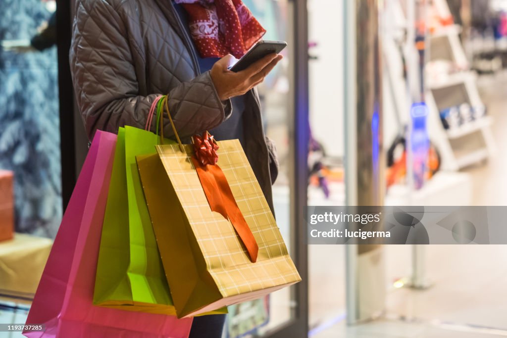 One people alone. A senior woman enjoys evening shopping while taking advantage of offers and discounts. On his arm a lot of shopping bags while she looks at her cell phone