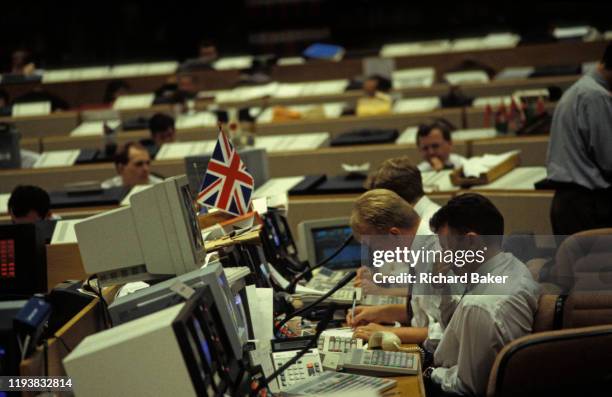 Currency banker with the British Union Jack above his desk, rubs tired eyes while working in front of 90s computers in the currency trading floor of...