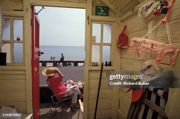 Seen from the inside of her beach hut that looks out to the promenade and sea, a lady enjoys summer sunshine while holding on to her hat while others...