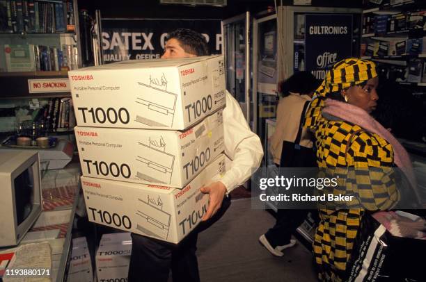 Shop assistant carries three boxes of Toshiba T1000 Portable Personal Computer laptops in an electronics and tech shop on the Tottenham Court Road,...