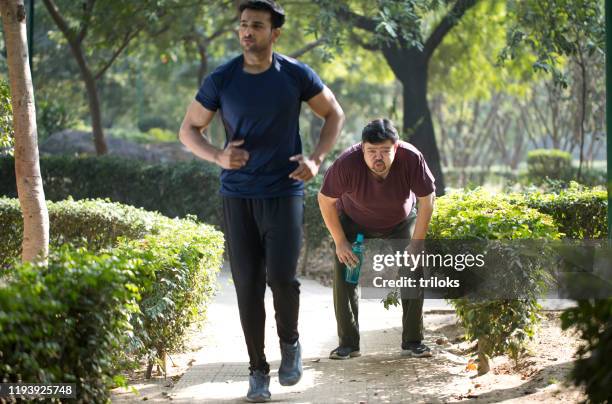 fitnesslehrer joggen, während erschöpfter dicker mann eine pause im park macht - atemnot stock-fotos und bilder