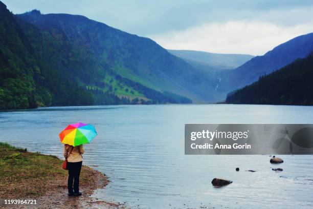 rear view of woman under rainbow umbrella standing by upper lake in wicklow mountains national park by glendalough, stormy spring skies - ireland rainbow stock pictures, royalty-free photos & images