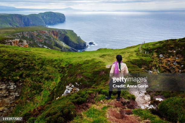 rear view of woman looking at steep cliffs of horn head and north atlantic ocean on overcast day in county donegal, ireland - condado de donegal fotografías e imágenes de stock