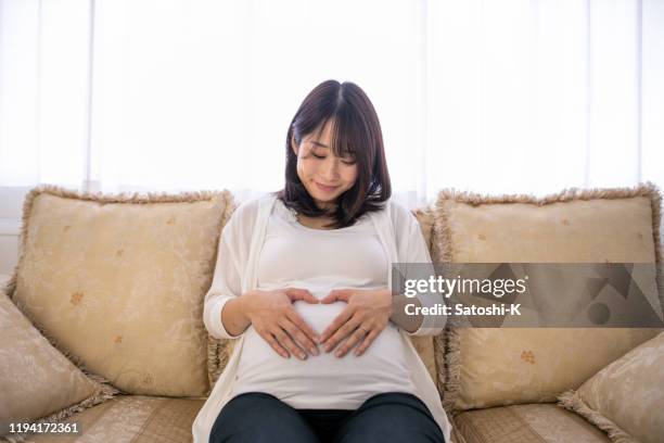 schwangere frau sitzt auf der couch und macht herzform mit ihren händen auf ihrem bauch - asian woman pregnant stock-fotos und bilder