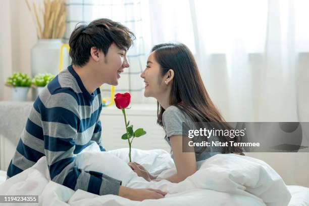 young asian couple in bedroom is sitting on bed with red rose at  home in bedroom.  enjoying spending time together. celebrating saint valentines day. - saint valentin stock pictures, royalty-free photos & images