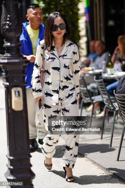 Guest wears earrings, sunglasses, a necklace, a lustrous pajamas style suit with black sharks pattern, black heeled sandals, a pink studded and...
